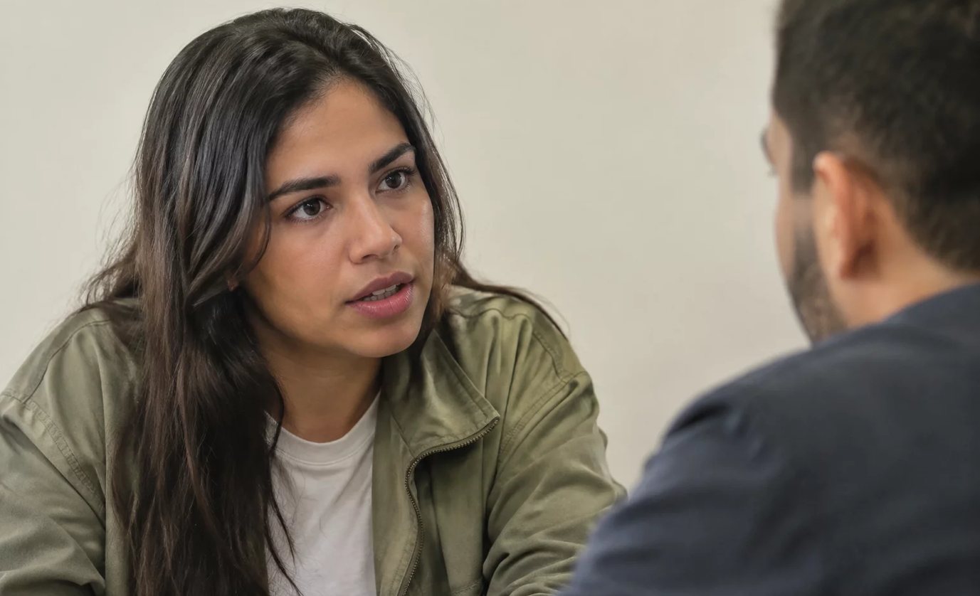 Três participantes conversam ao redor de uma mesa comprida numa sala clara de janelas altas